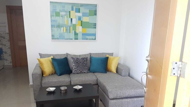 Un sofá seccional gris con cojines amarillos y azules, una mesa de centro negra y un cuadro abstracto moderno en la pared.