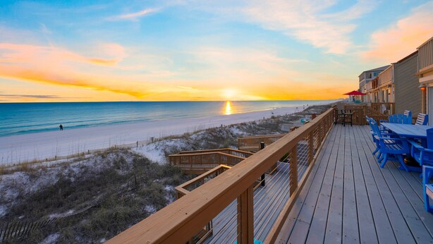 Un pontile di legno con sedie e tavoli blu che si affaccia su una spiaggia al tramonto.