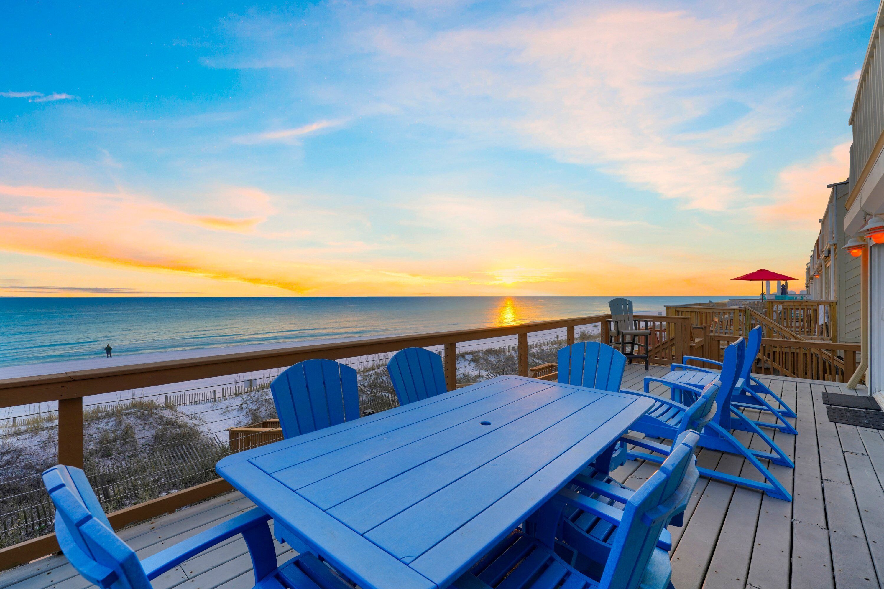 Een houten terras met blauwe stoelen en een tafel, met uitzicht op de oceaan tijdens de zonsondergang.