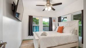 Ein Schlafzimmer mit Bett, Deckenventilator, Fenster mit Vorhängen und Blick ins Grüne.