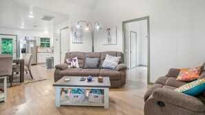 Ein geräumiger Wohnbereich mit braunem Sofa, Couchtisch und Esstisch. An den Wänden hängen gerahmte Bilder und im Hintergrund ist eine Küche zu sehen.