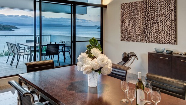 Un comedor con vista al mar, un jarrón de flores blancas y una botella de vino sobre la mesa.
