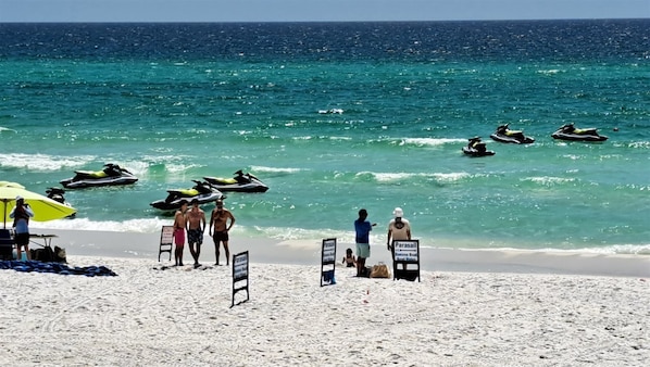 A beach with jet skis, a parasail sign, and people on the sand.