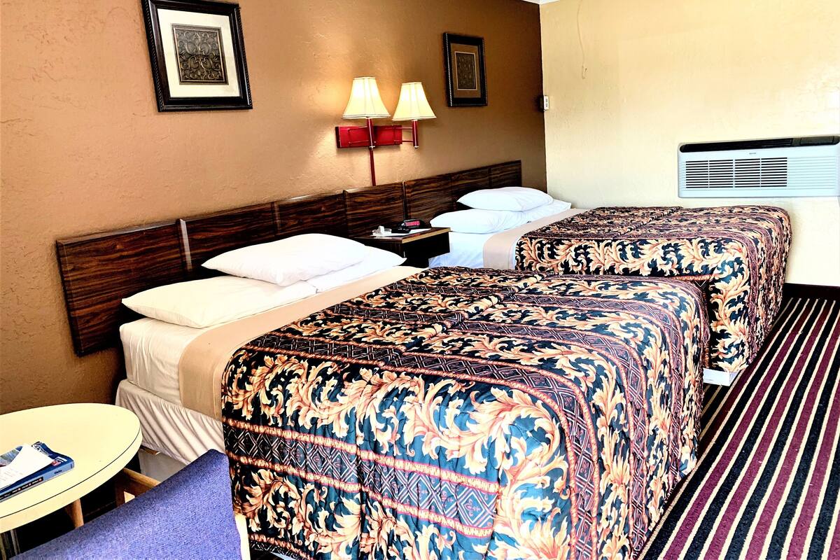 Une chambre d’hôtel avec deux lits, une table de chevet, une lampe, un tableau encadré et une fenêtre avec un appareil de climatisation.