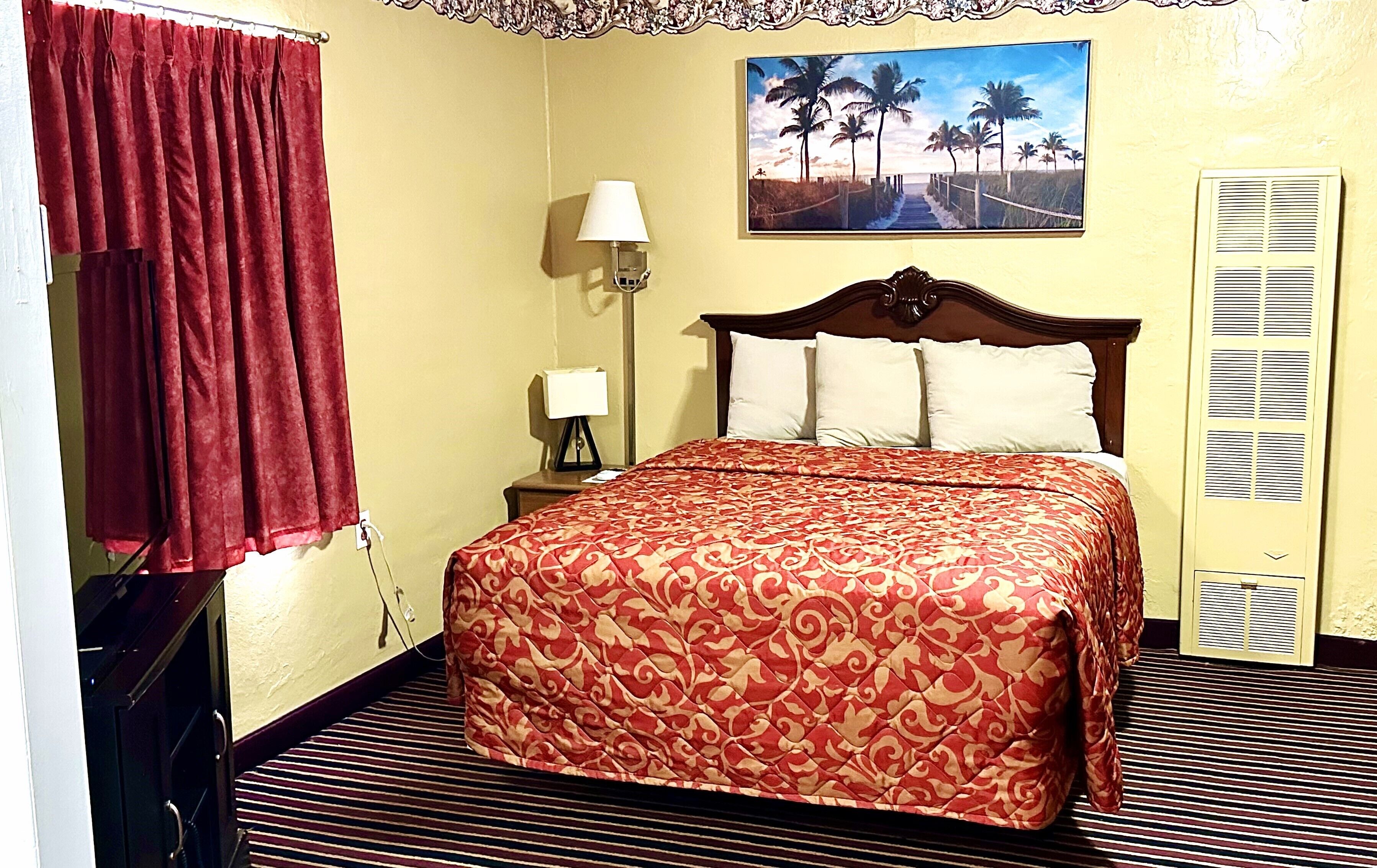 Habitación de hotel con cama, mesita de noche, lámpara, un cuadro de una playa y una ventana con cortinas.