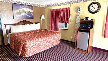 A hotel room with a bed, a wall clock, an air conditioning unit, a microwave, and a small refrigerator.