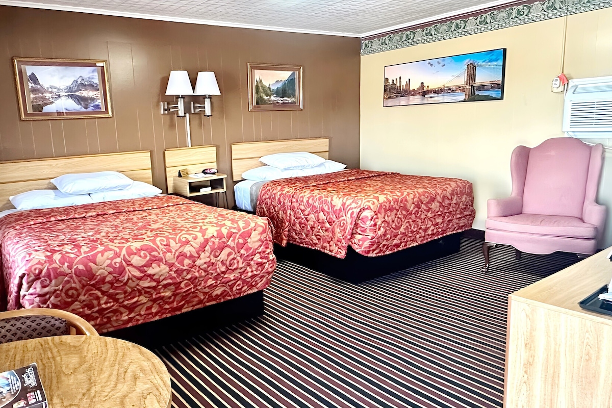 A hotel room with two beds, a wooden headboard, a pink armchair, a desk, and a patterned carpet.