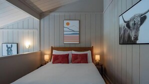 A neatly made bed with white and red bedding, a wooden headboard, and a wall-mounted artwork depicting a landscape with a sun and mountains.