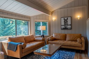 Un salon avec deux canapés en cuir, une table basse en verre et une lampe. Il y a des fenêtres donnant sur des arbres, et un tableau encadré au mur.