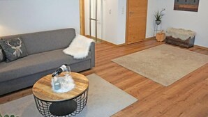 Olohuoneessa on harmaa sohva, pyöreä puinen sohvapöytä, jonka päällä on valkoinen, pehmeä huopa, beigenvärinen matto ja puulattia.