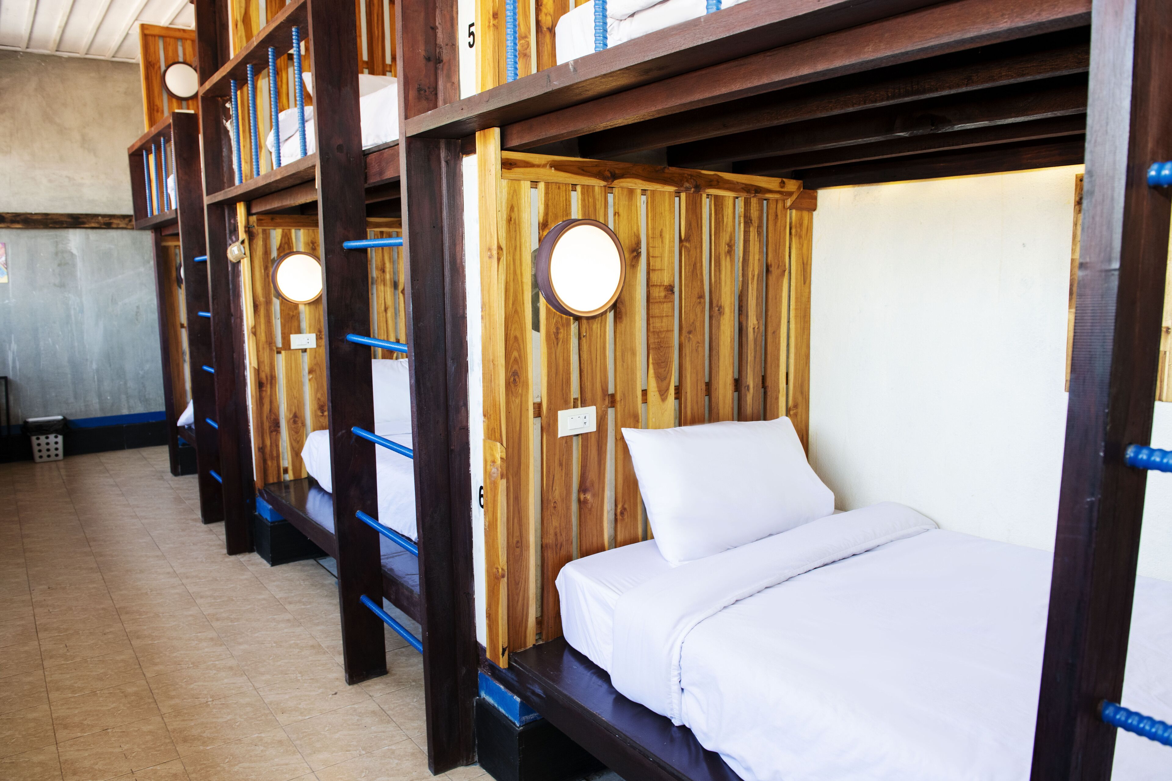 A row of bunk beds with wooden frames and blue railings, each with a round wall-mounted light.