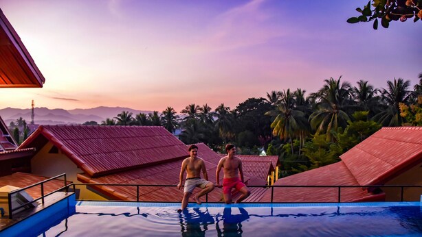 A swimming pool with two people standing at the edge, surrounded by red-roofed buildings and palm trees. - Mad Monkey Luang Prabang (Luang Prabang)