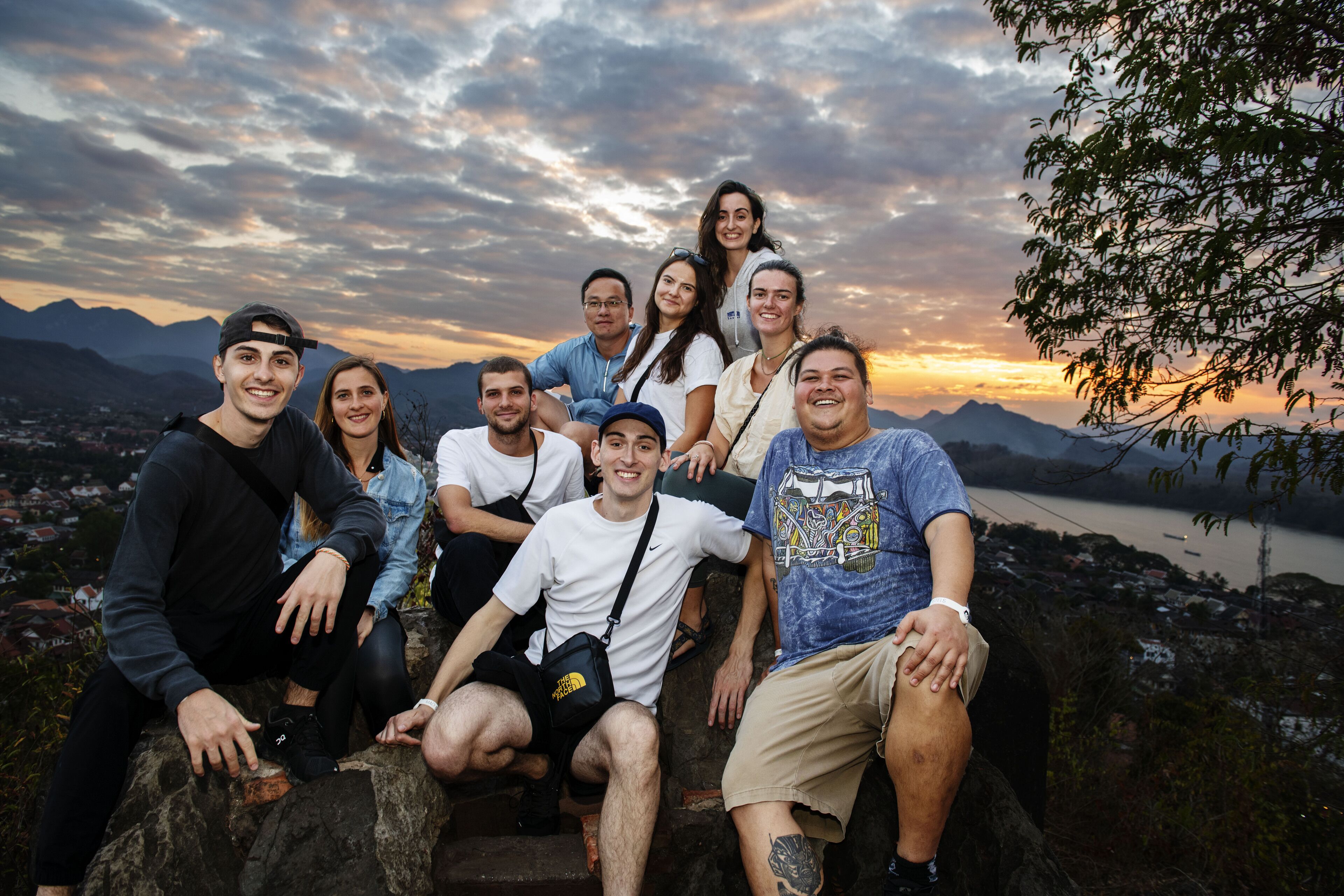A group of people sitting on a rock with a scenic view of mountains and a river in the background.