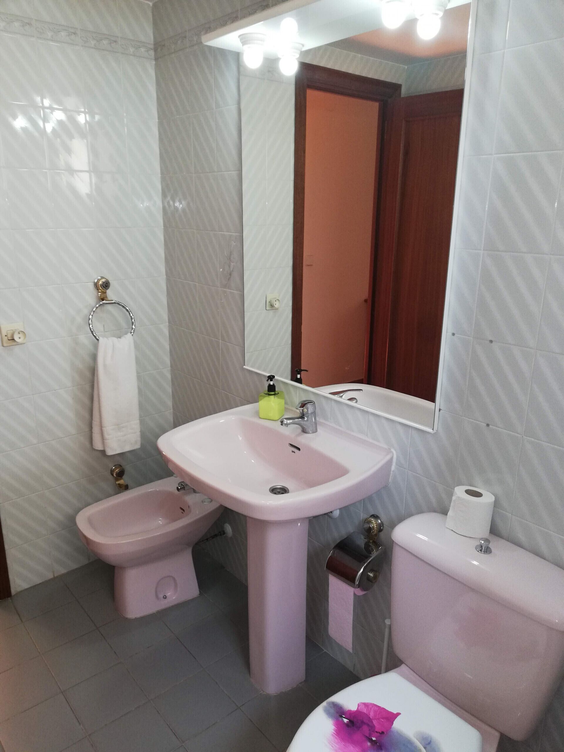 Et badeværelse med en lyserød vask, bidet, toilet og et spejl med lys over.