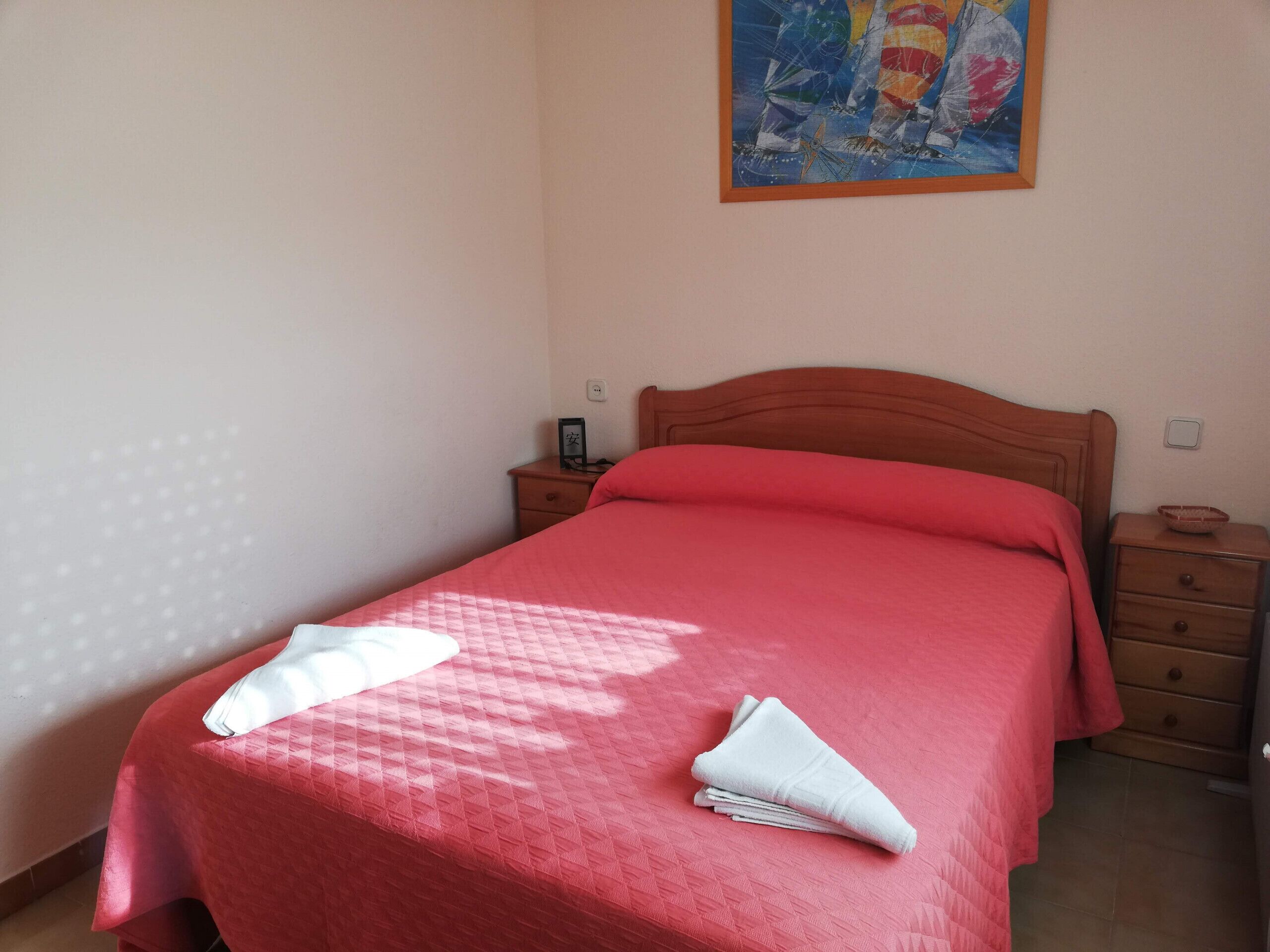 A neatly made bed with a red bedspread and pillows, a wooden headboard, a small bedside table with a lamp, and a painting on the wall.