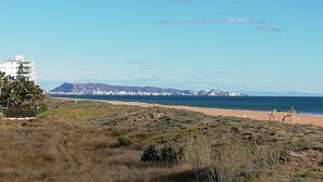 A coastal landscape with a beach, a mountain range in the background, and a white building with balconies. - La Marina I 4º Solo Familias! (Xeraco Playa)