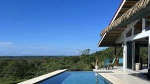 Une piscine à débordement moderne, avec une eau d’un bleu cristallin, entourée d’une terrasse en pierre et d’une structure surmontée d’un toit de chaume.