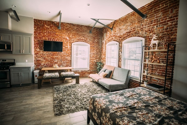 Un espacio habitable compacto con pared de ladrillo, un sofá, una cama, un televisor y una zona de cocina.