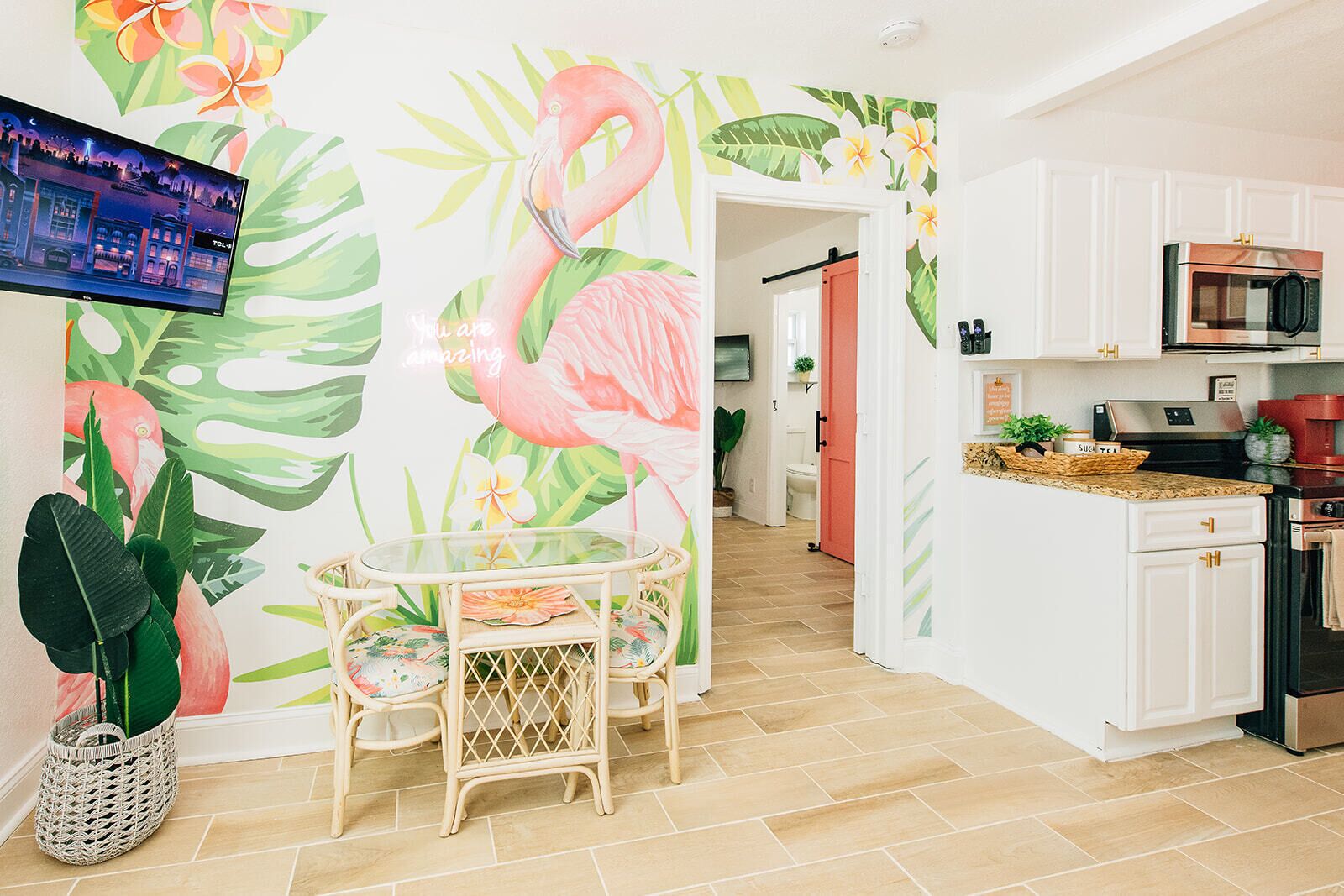 A modern kitchen with white cabinets, a microwave, and a coffee maker. There is a dining area with a glass table and chairs, and a wall with a tropical mural.