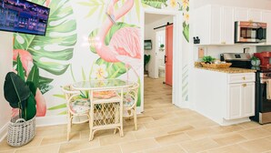 A modern kitchen with white cabinets, a microwave, and a coffee maker. There is a dining area with a glass table and chairs, and a wall with a tropical mural.