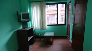 A small, green-walled room with a television on a shelf, a window with curtains, a wooden bench, and a door. - Green House Lodge (Kathmandu)