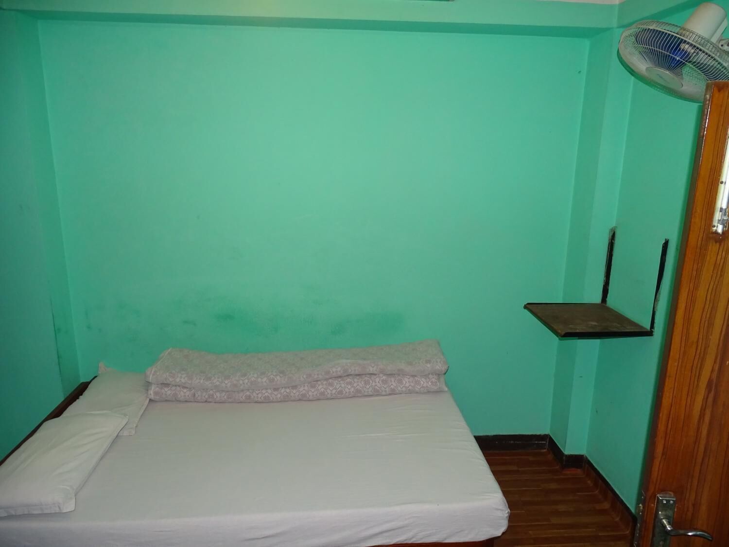 Una habitación pequeña con una cama individual, piso de madera, pared verde y un ventilador de pie.