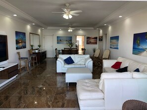 Uma sala de estar ampla, com um sofá branco, um espaço para refeições e uma cozinha ao fundo.