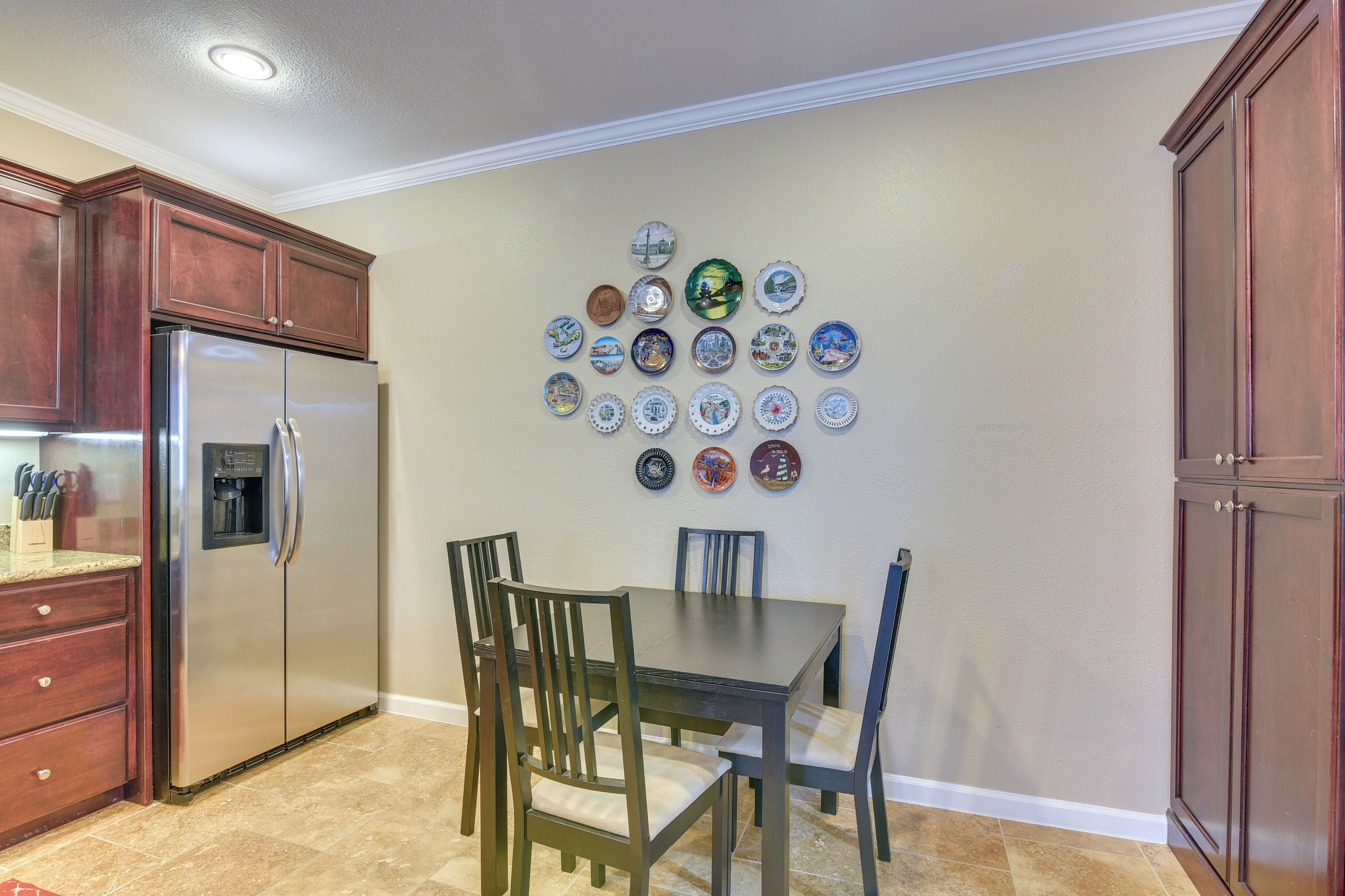 A kitchen with a stainless steel refrigerator, wooden cabinets, a dining table with chairs, and a wall decorated with various plates.