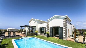 Una casa moderna de dos pisos con piscina, mobiliario exterior y vista al océano.