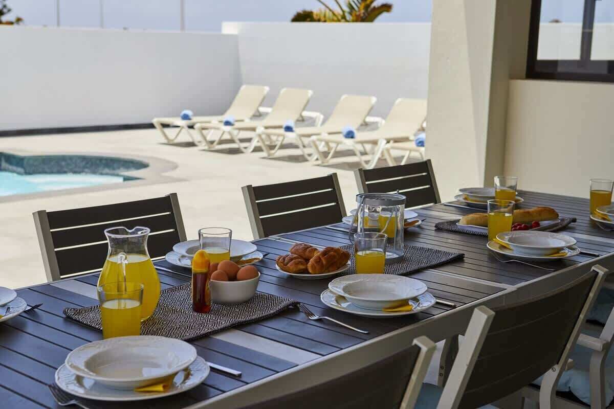 A dining table set with breakfast items, a pitcher of orange juice, and a bowl of eggs, overlooking a pool with lounge chairs.