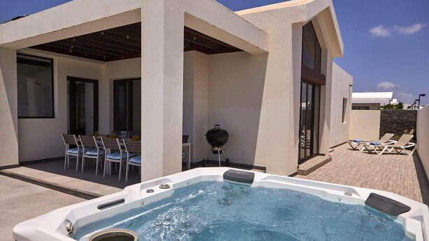 Un área moderna de piscina exterior con un jacuzzi, una parrilla y un patio con sillas y tumbonas.