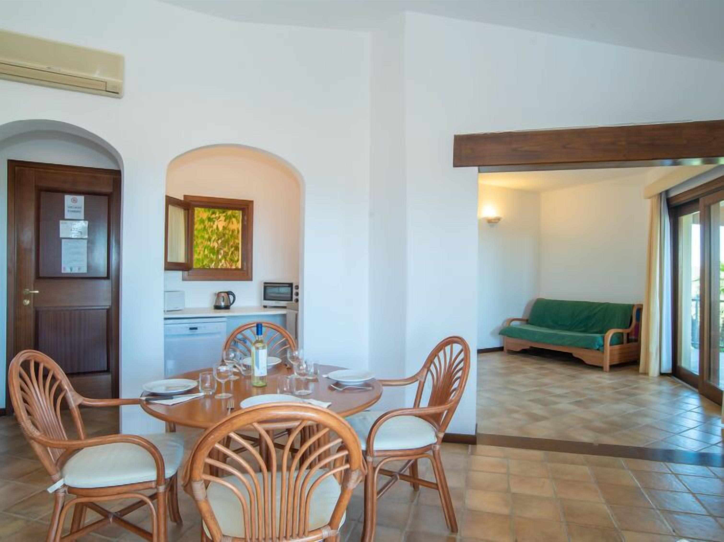 Un comedor con una mesa redonda y cuatro sillas de mimbre, una kitchenette con microondas y hervidor, y un sofá verde en una sala de estar con vistas a través de una puerta de vidrio.