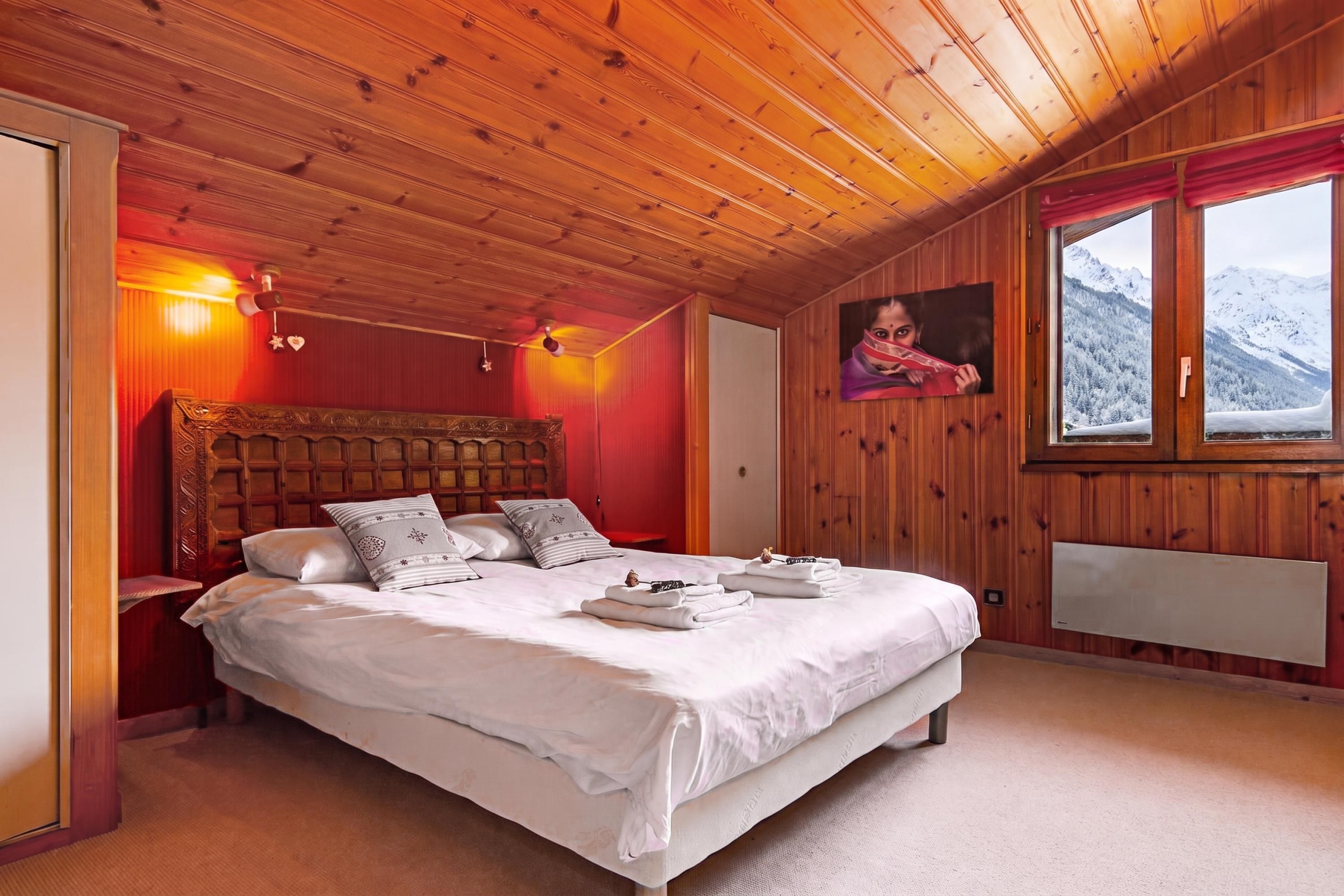 Une chambre avec un plafond en bois, un lit recouvert de draps blancs, une table de chevet, un radiateur fixé au mur et une fenêtre donnant sur la montagne.