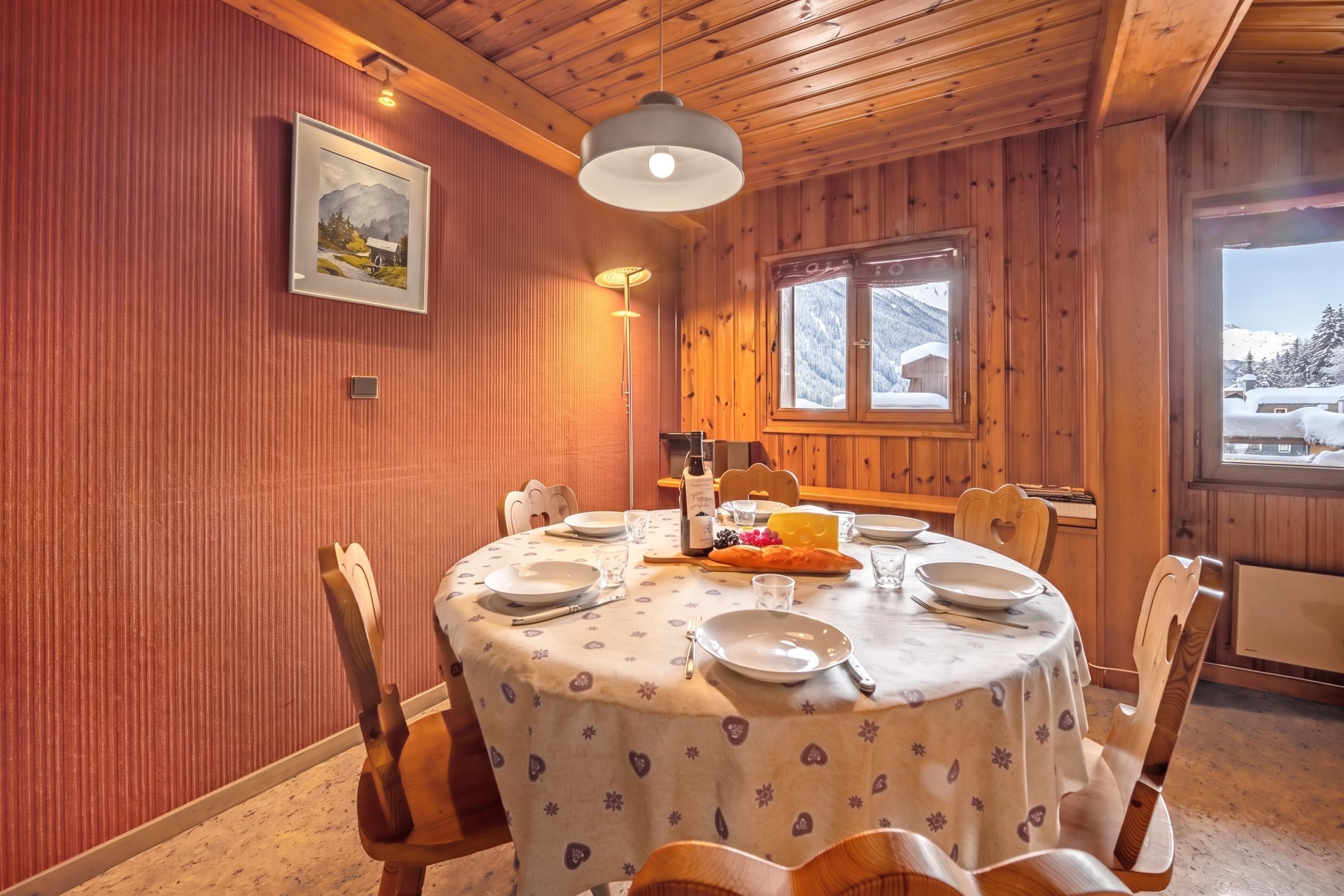 Ein Essbereich mit einem runden Tisch, gedeckt für vier Personen, Holzstühlen, einem Fenster mit Blick auf schneebedeckte Berge und einem gerahmten Bild an der Wand.