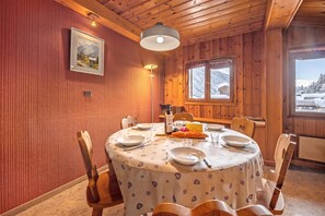 A dining area with a round table set for four, wooden chairs, a window with a view of snow-covered mountains, and a framed picture on the wall.