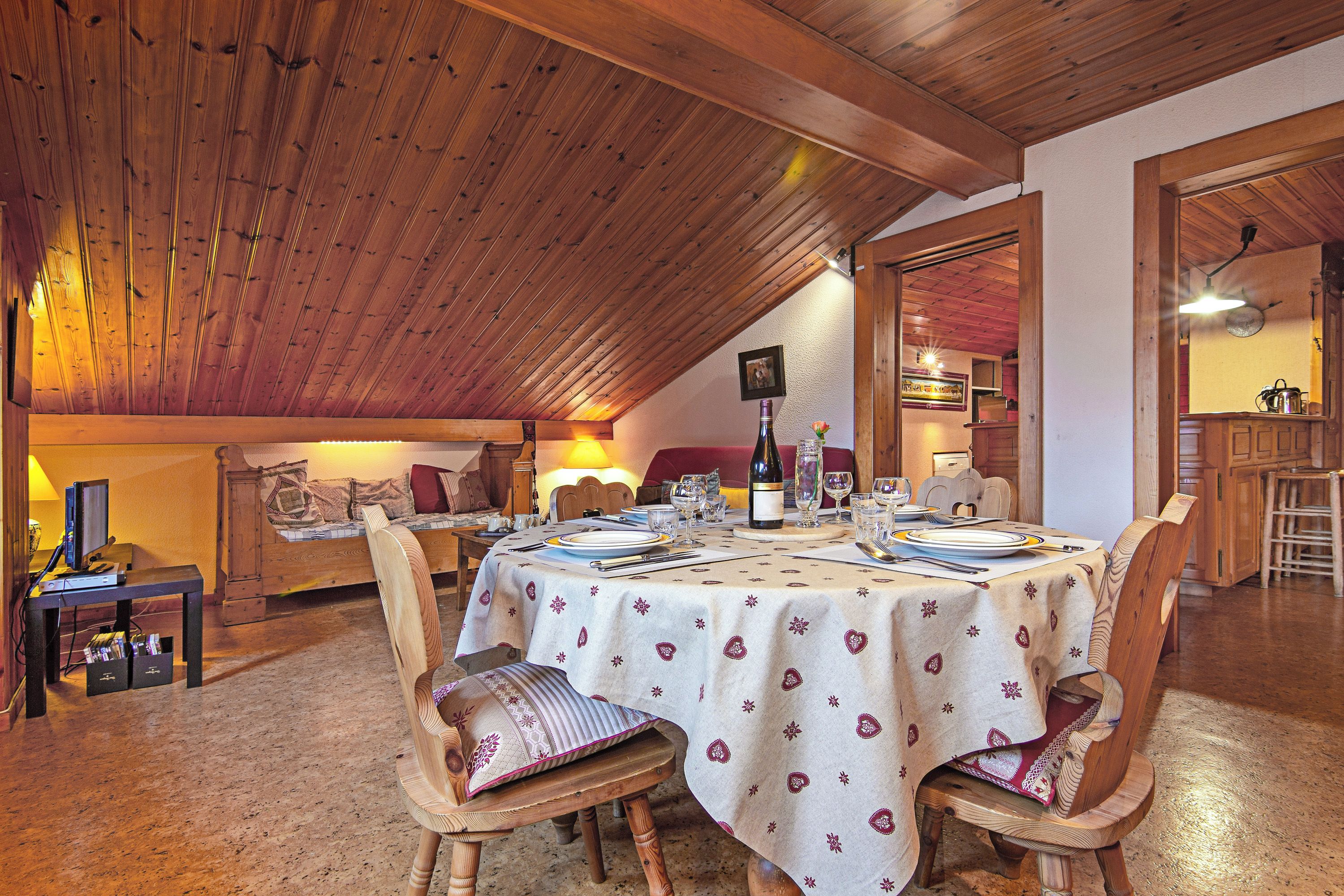 Un espace repas avec une table dressée pour quatre personnes, des chaises en bois et un haut plafond également en bois.