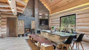 A log cabin interior with a stone fireplace, a glass dining table, and a large window offering a view of the outdoors.