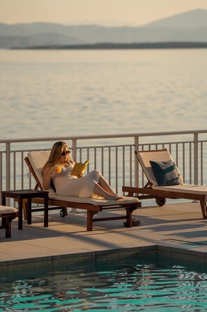 A person relaxing on a lounge chair by a pool with a view of the water and mountains in the background.