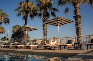 Un área junto a la piscina con sillones de descanso y sombrillas, rodeada de palmeras.