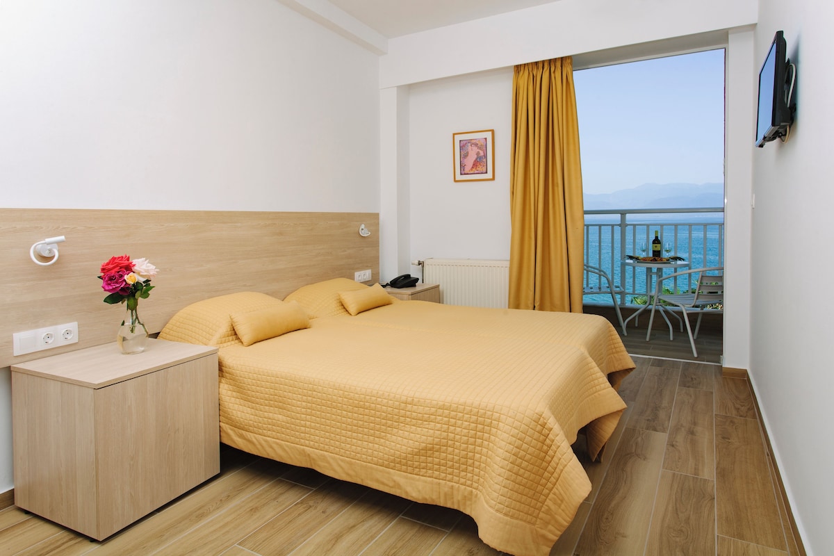 Habitación de hotel con cama, mesita de noche, una pequeña mesa con una botella y vista al mar desde el balcón.