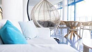 A modern hotel room with a hanging chair, a sofa with blue pillows, and wicker chairs around a small table. - Sissy (Kamena Vourla)