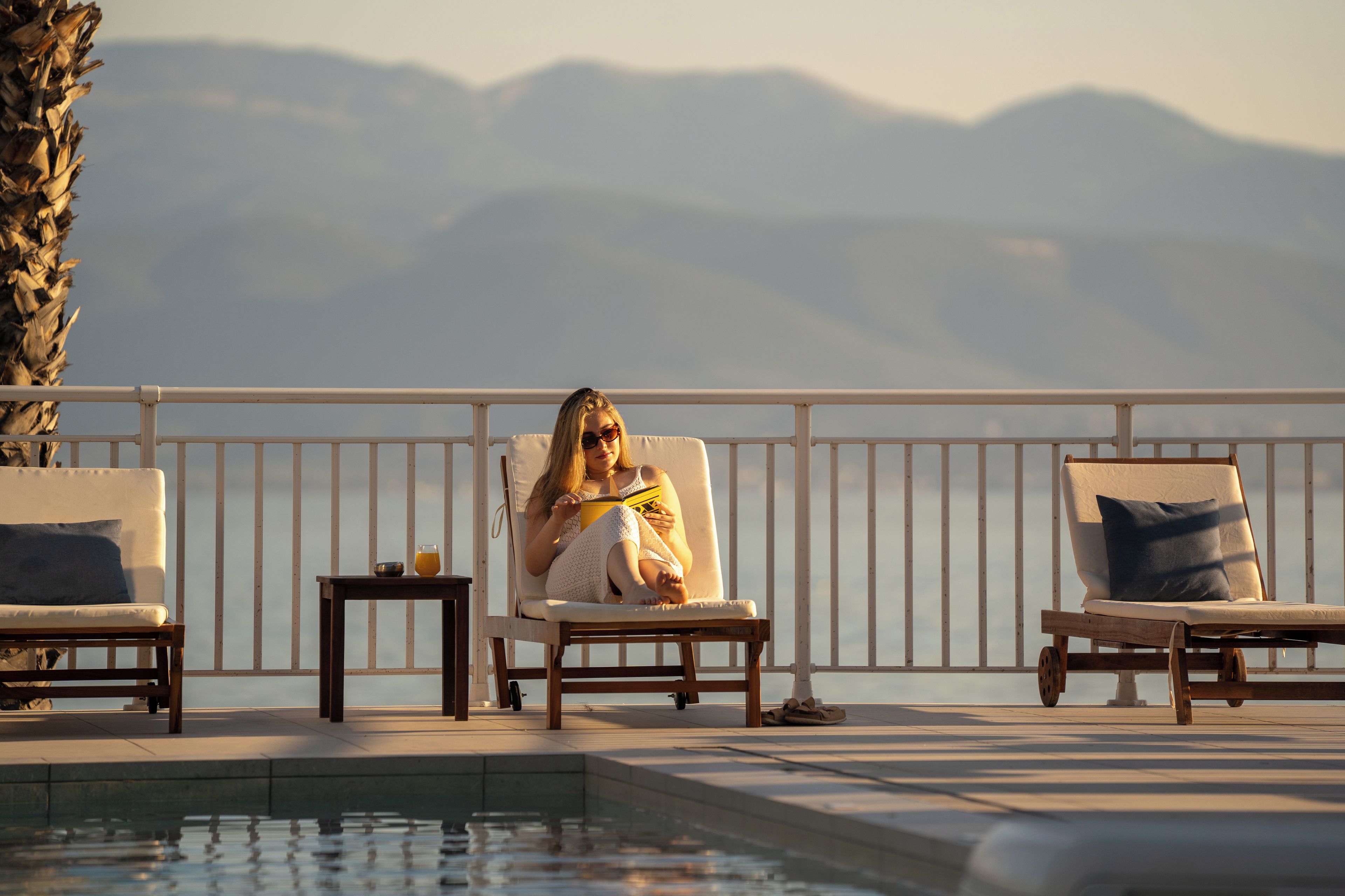 A poolside area with lounge chairs, a table with a drink, and a view of mountains.
