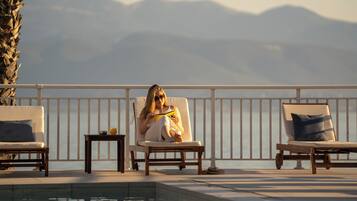A poolside area with lounge chairs, a table with a drink, and a view of mountains.