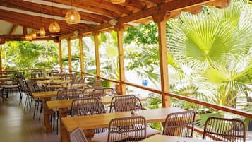 A covered outdoor dining area with wooden tables and chairs, surrounded by lush greenery.