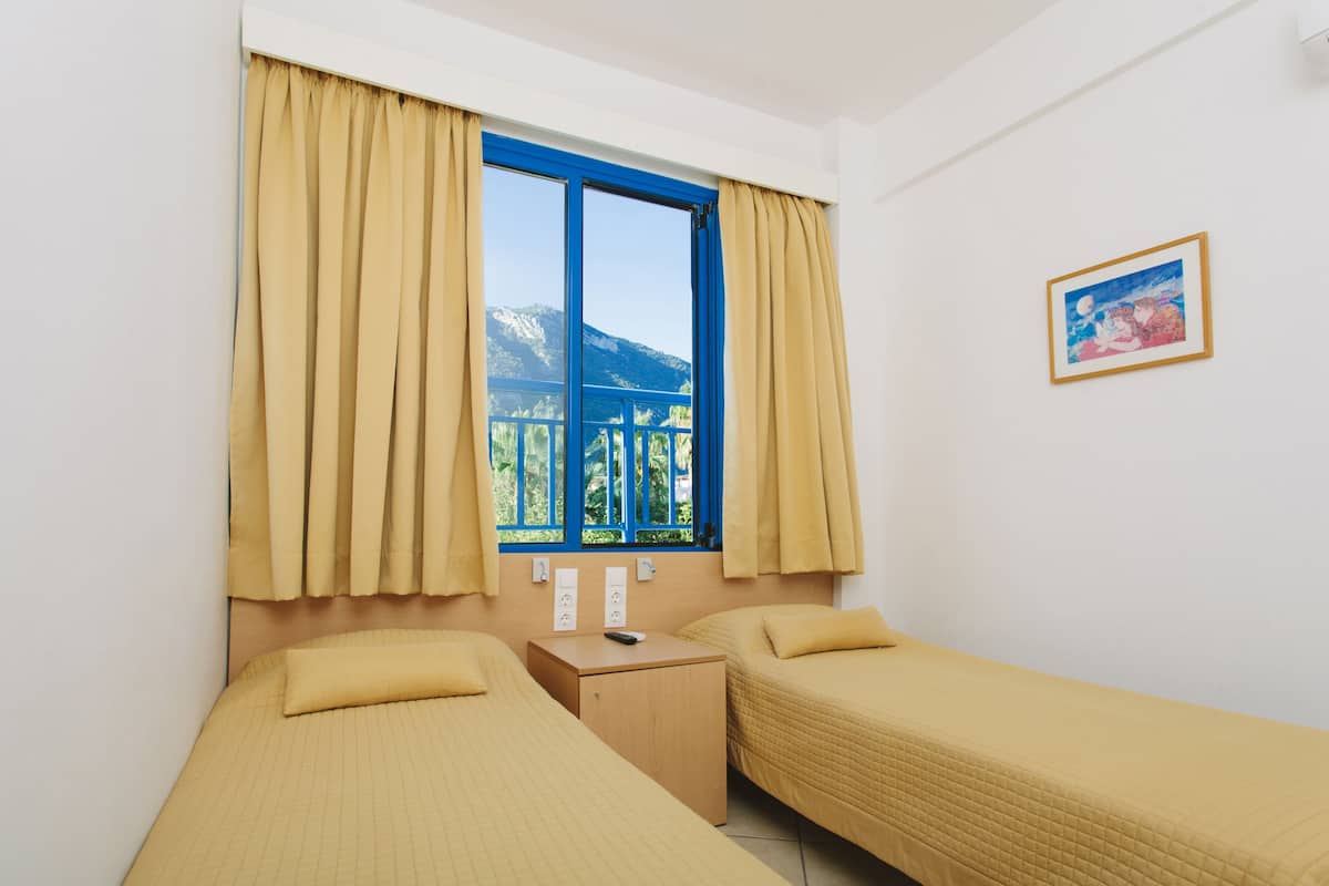 A room with two beds, a window with curtains, a small bedside table, a painting on the wall, and a view of mountains through the window.