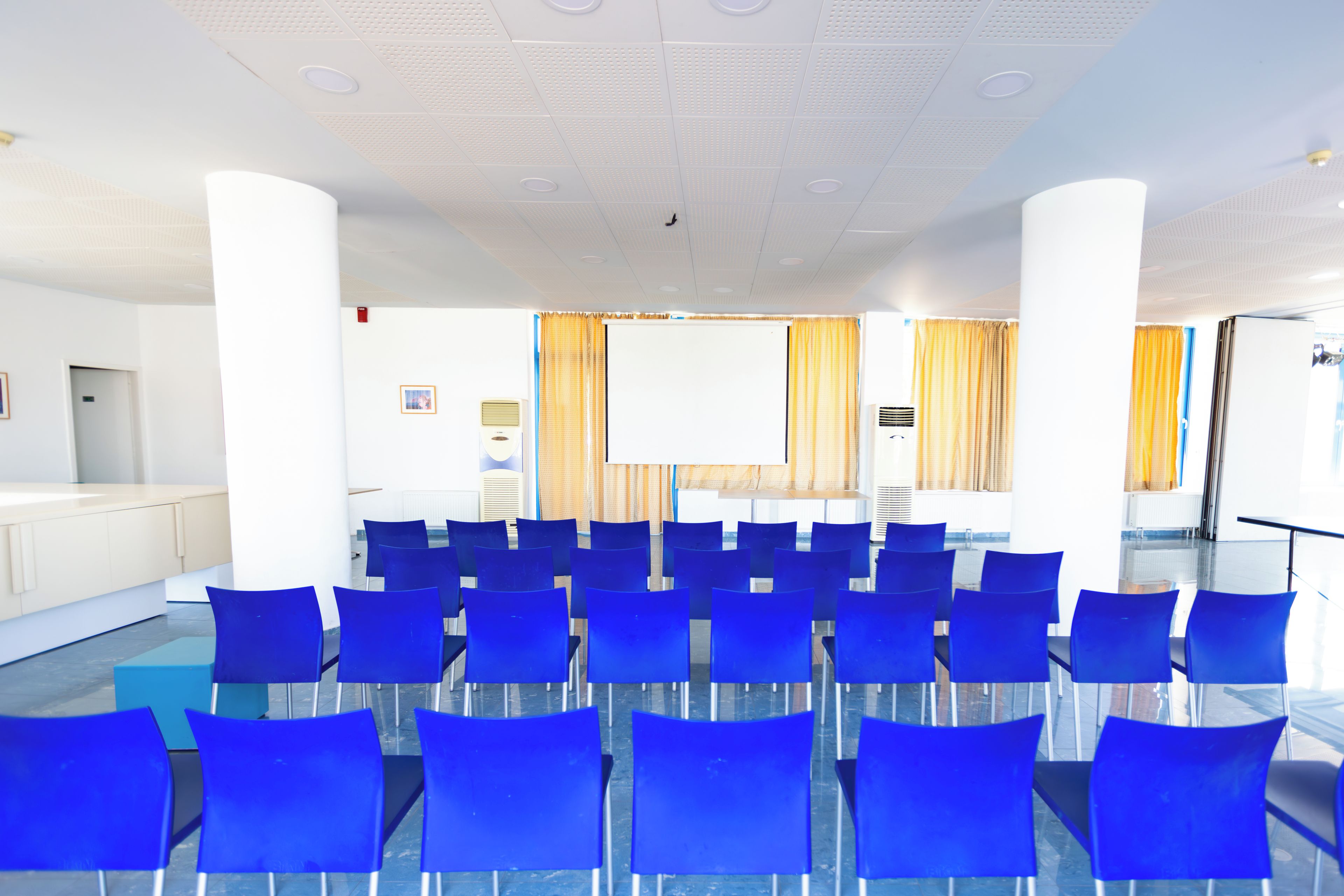 A conference room with rows of blue chairs, a large white screen, and a podium.