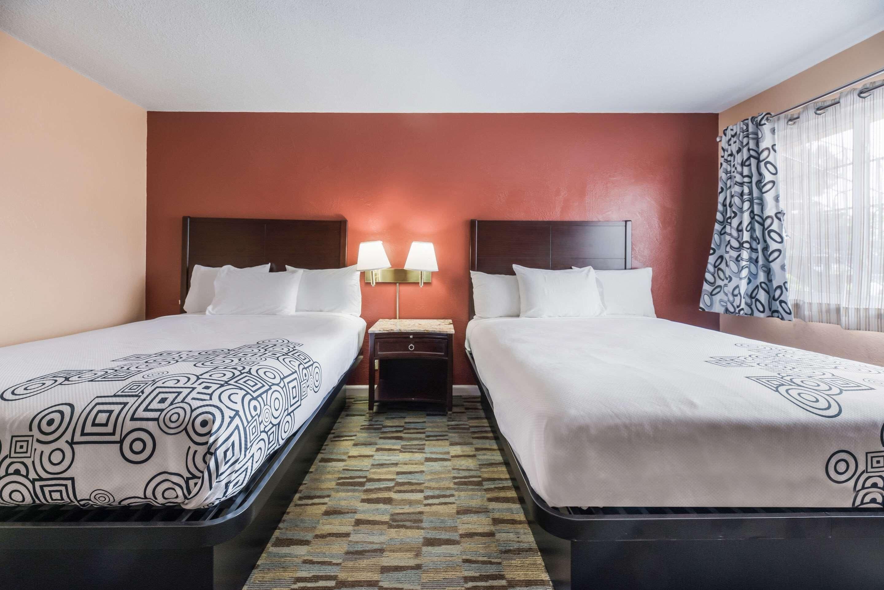 A hotel room with two beds, a patterned bedspread, a small nightstand with lamps, and a patterned rug.