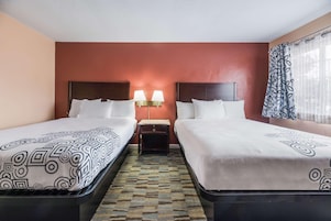 Habitación de hotel con dos camas, una colcha estampada, una mesita de noche con lámparas y una alfombra con diseño.