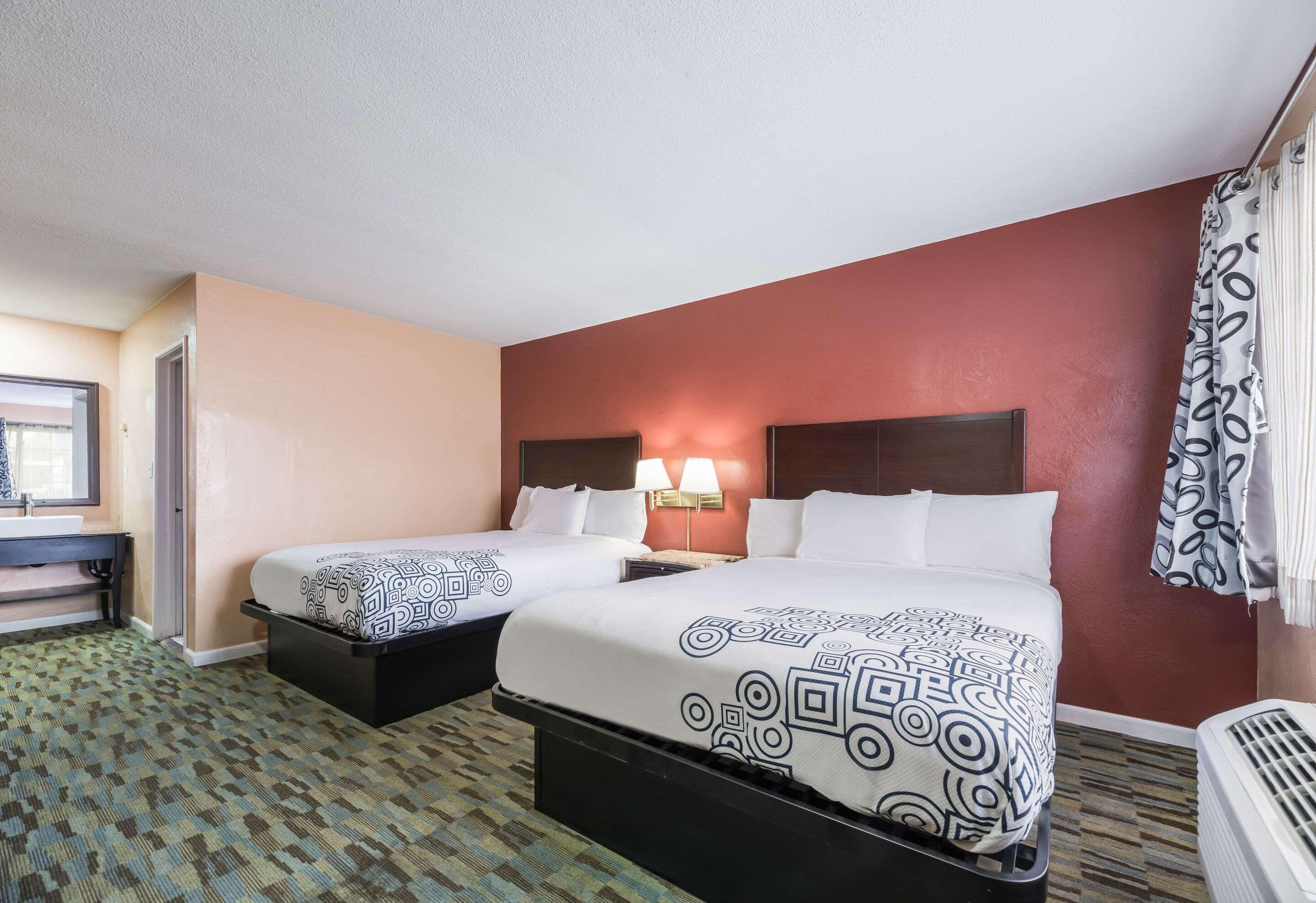 A hotel room with two beds, a desk, a chair, a lamp, and a window.