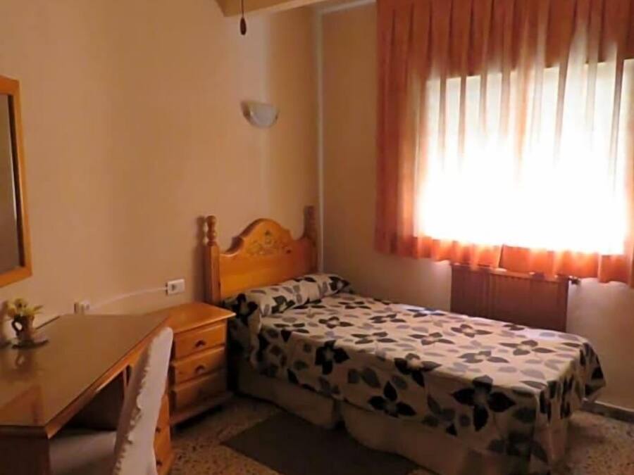 Un dormitorio con cama, escritorio, silla, ventilador de techo y ventana con cortinas.